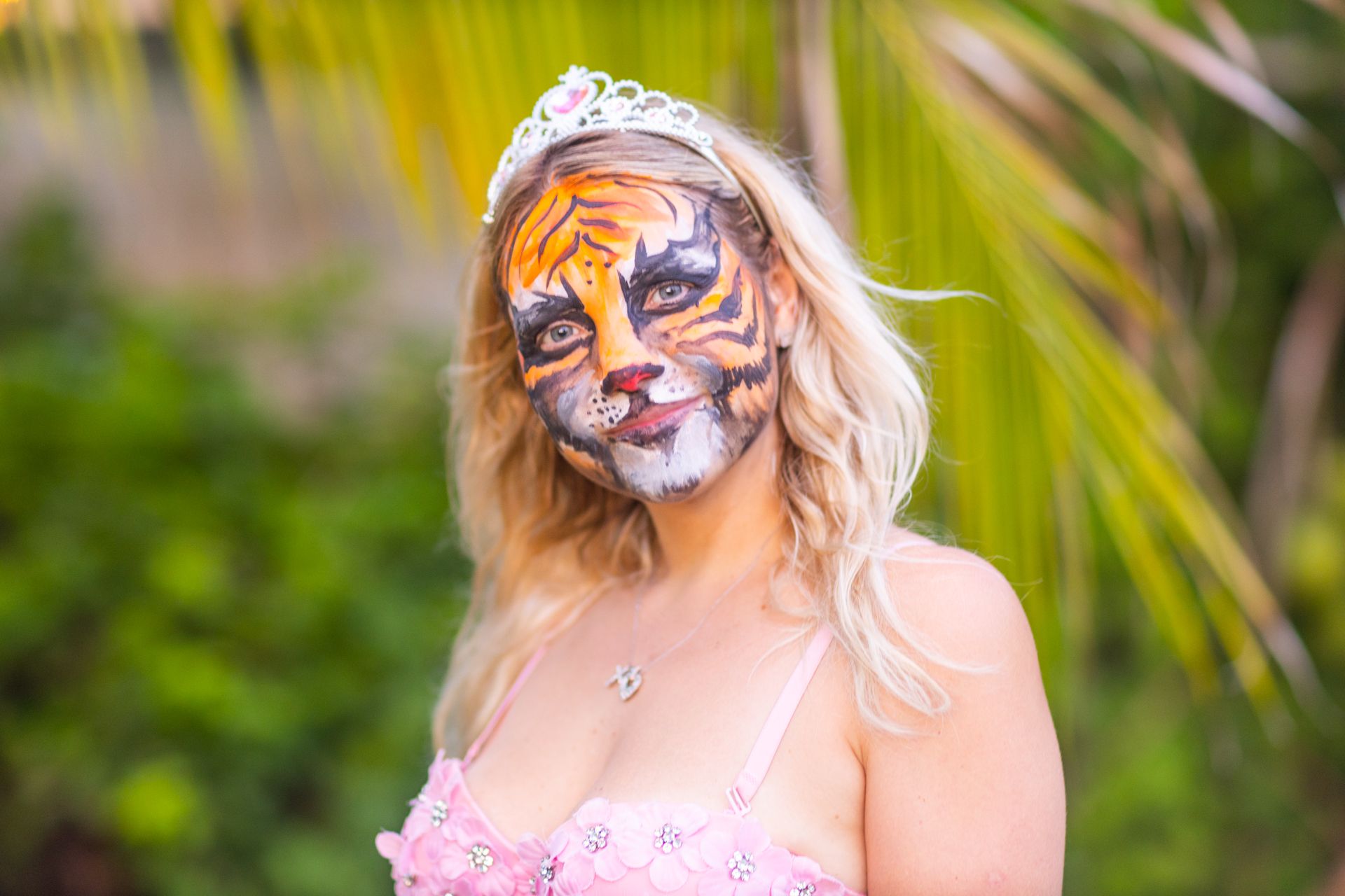 A young woman with a painted tiger face and a silver tiara, palm leaves behind her